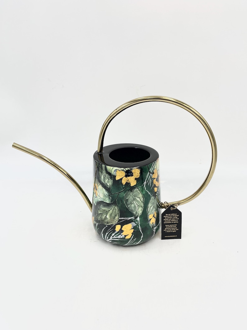 May include: A black watering can with a gold handle and a green and gold floral pattern. The can has a spout and a small tag attached to the handle.