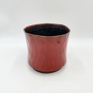May include: A red ceramic planter with a black glazed interior. The planter has a slightly rounded shape and a textured surface.