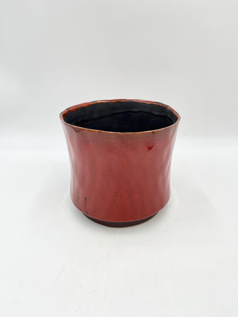 May include: A red ceramic planter with a black glazed interior. The planter has a slightly rounded shape and a textured surface.