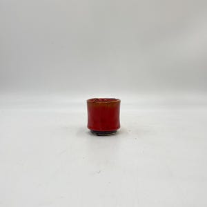 May include: A small, red ceramic cup with a slightly flared rim. The cup has a dark brown glaze on the bottom.
