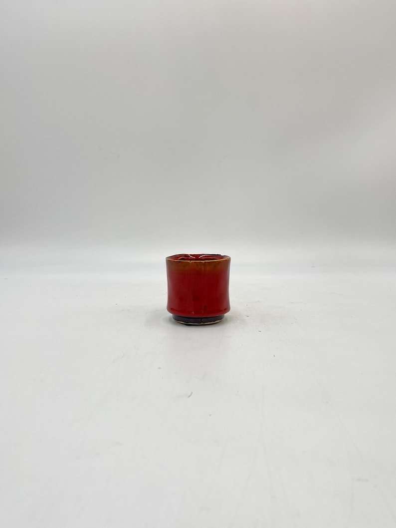 May include: A small, red ceramic cup with a slightly flared rim. The cup has a dark brown glaze on the bottom.