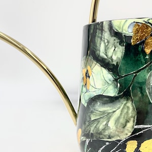 May include: A gold watering can with a black and green floral design. The design features large green leaves and small yellow flowers with gold accents.