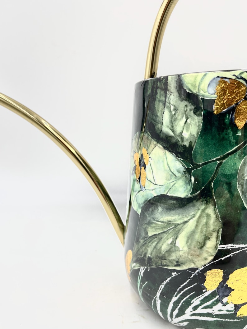 May include: A gold watering can with a black and green floral design. The design features large green leaves and small yellow flowers with gold accents.