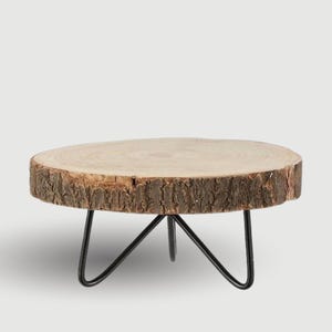 May include: A wooden cake stand with a natural bark edge. The round wooden top is supported by three black metal legs. The stand is ideal for displaying cakes or other treats. The diameter is approximately 25 cm.