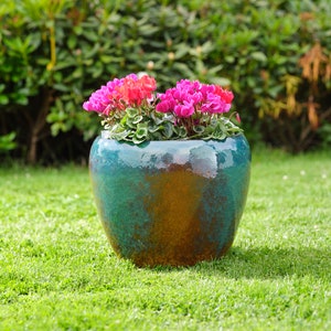 May include: A green glazed ceramic planter with pink and red flowers. The planter is sitting on a green grassy lawn.