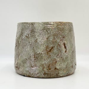 May include: A handmade ceramic plant pot with a textured, mottled finish. The pot displays a mix of earthy colours, including greens, browns, and greys. The vessel has a wide opening and a gently sloping form, suitable for various plants.