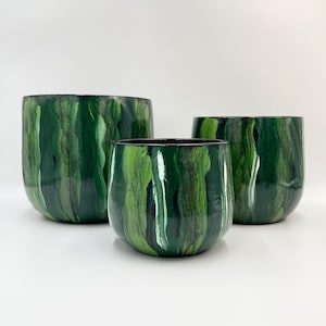 May include: Three green and black painted ceramic planters of varying sizes. The planters have a rounded shape and a glossy finish. The green paint is streaked with black and white, creating a marbled effect. The interior of the planters is black.