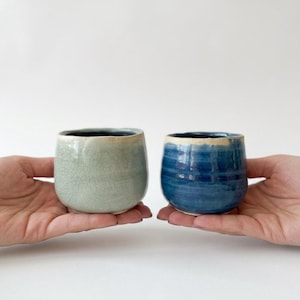 May include: Two small, rounded ceramic cups with a smooth, glossy finish. The cup on the left is a pale, crackled mint green, while the cup on the right is a deep, vibrant cobalt blue with subtle horizontal ridges along the exterior surface.