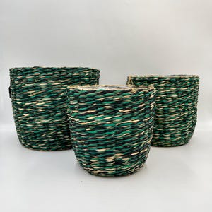 May include: Three woven baskets of different sizes. The cylindrical baskets have a textured surface. The weave features shades of green, black, and natural beige. These baskets are suitable for storage or decorative purposes.