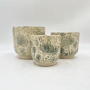 May include: Three ceramic plant pots with a white background and green floral designs. The pots are different sizes and are arranged in a row from largest to smallest.