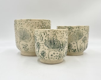 May include: Three ceramic planters with a white background and green floral designs. The planters are different sizes and are arranged in a row from largest to smallest.