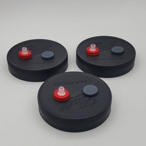 Puede incluir: Tres tapas negras para tarros con componentes rojos y grises. Cada tapa tiene la palabra "Ball" y "LEAK-PROOF" impresa. Las tapas están diseñadas para enlatar y conservar.
