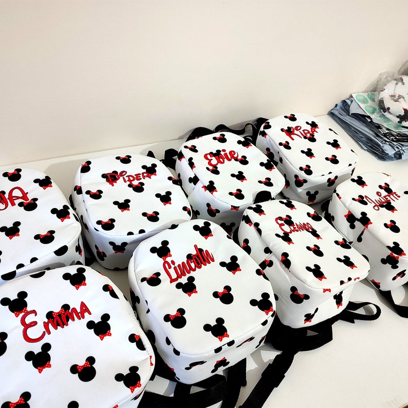 May include: Nine white backpacks with black straps and red bows. Each backpack has a different name embroidered on it in red: Piper, Evie, Lincoln, Emma, Ava, Juliette, Liam, and two others.