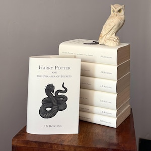 Op de afbeelding: Een stapel Harry Potter-boeken van J.K. Rowling, met een witte kaft en een uilbeeldje. Het voorste boek is "Harry Potter en de Geheime Kamer" met een zwarte slangenillustratie. De boeken staan op een houten oppervlak.