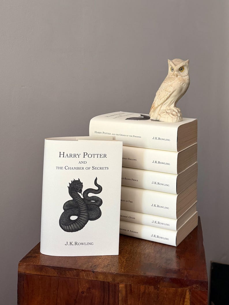 May include: A stack of Harry Potter books by J.K. Rowling, with a white cover and an owl figurine. The book in front is "Harry Potter and the Chamber of Secrets" with a black serpent illustration. The books are on a wooden surface.