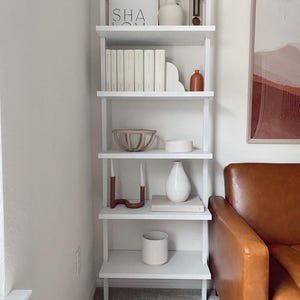 May include: White ladder shelf with five shelves, decorated with books, candles, vases, and a wooden bowl. The shelf is in a corner of a room with a brown leather couch in the background.