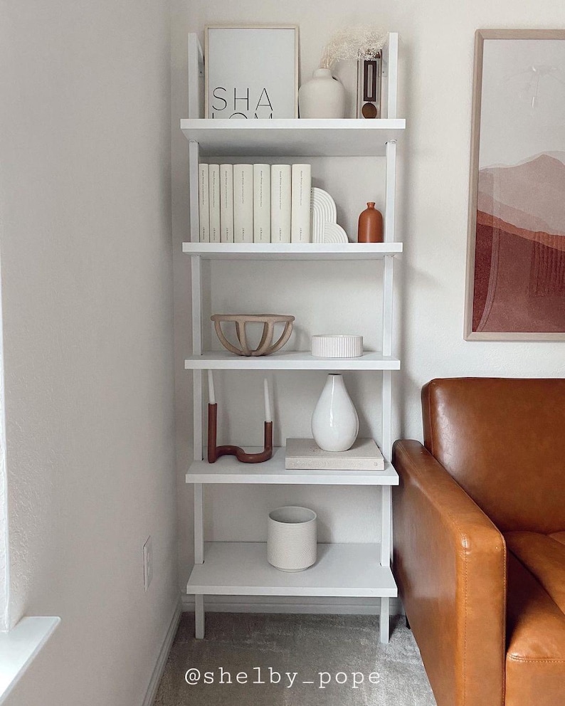 May include: White ladder shelf with five shelves, decorated with books, candles, vases, and a wooden bowl. The shelf is in a corner of a room with a brown leather couch in the background.