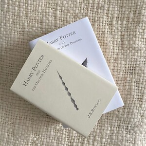 May include: Two hardcover books from the Harry Potter series. The top book is titled "Harry Potter and the Order of the Phoenix." The bottom book is titled "Harry Potter and the Deathly Hallows" and features a wand design. Both books are on a textured, off-white surface.