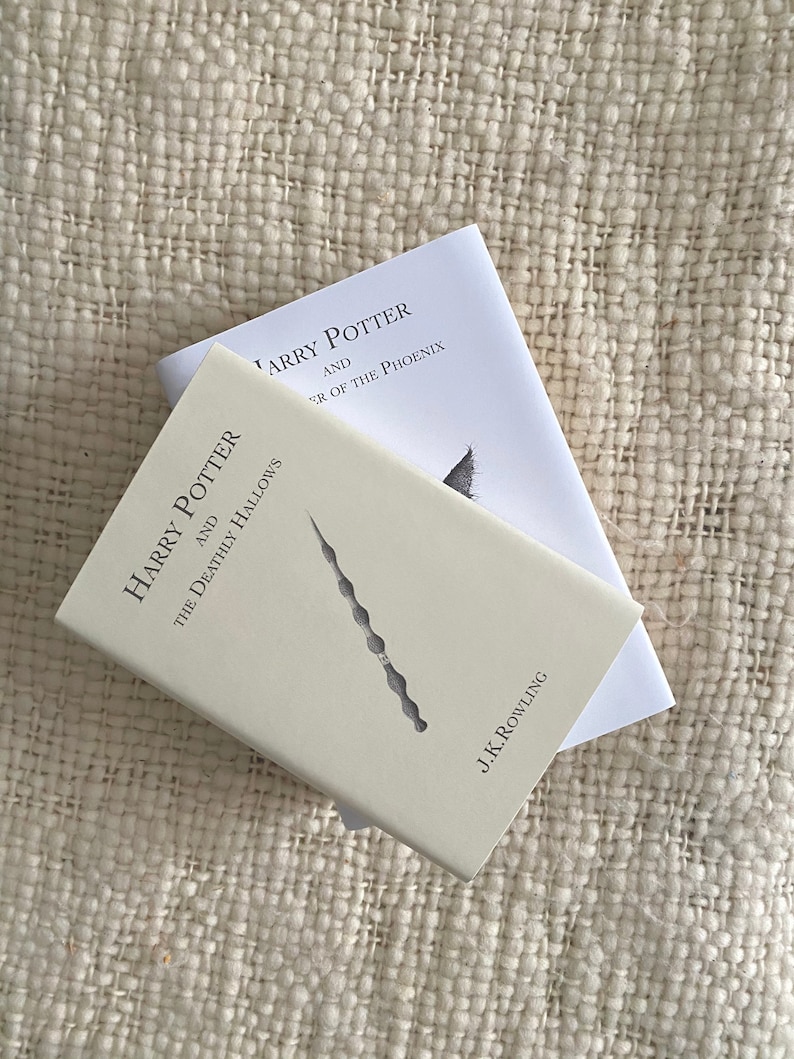 May include: Two hardcover books from the Harry Potter series. The top book is titled "Harry Potter and the Order of the Phoenix." The bottom book is titled "Harry Potter and the Deathly Hallows" and features a wand design. Both books are on a textured, off-white surface.
