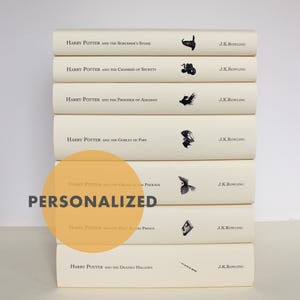 以下が含まれることがあります： ハリー・ポッターシリーズの7冊の本の積み重ね。各本はクリーム色の表紙と黒いイラストが特徴です。タイトルと著者であるJ.K.ローリングの名前が背表紙に印刷されています。「PERSONALIZED」という文字が画像に重ねられています。