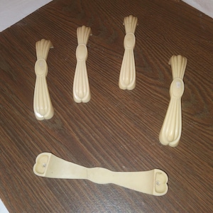 Op de afbeelding: Een verzameling van vijf crèmekleurige, tarwevormige kastknoppen en een bijpassende handgreep. De knoppen hebben een gedetailleerd ontwerp en de handgreep is gebogen met twee montagegaten. Geplaatst op een donkerbruin houten oppervlak.