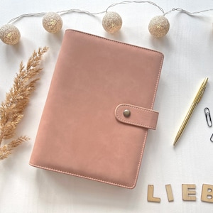 Könnte beinhalten: Ein blushfarbenes Leder-Notizbuch mit goldfarbenem Knopfverschluss. Das Notizbuch liegt auf einer weißen Oberfläche mit einer Lichterkette und einem getrockneten Pampasgras-Zweig. Das Wort "LIEBS" ist in Holzbuchstaben geschrieben.