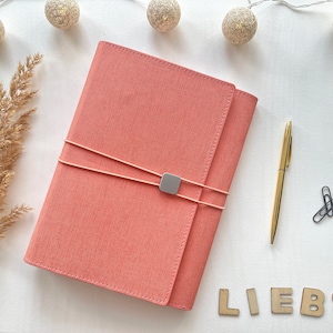 Könnte beinhalten: Ein korallenfarbenes, stoffbezogenes Notizbuch mit einem weißen Kordelverschluss und einem silbernen Metallakzent. Das Notizbuch liegt auf einer weißen Oberfläche mit einem Zweig getrockneten Pampasgrases, einem goldenen Stift und dem Wort "LIEBS" in Holzbuchstaben.