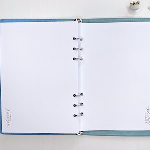 Könnte beinhalten: Ein blaues und weißes Notizbuch mit einem gepunkteten Muster und einem silbernen Binder. Das Notizbuch ist auf einer leeren Seite geöffnet, auf der die Worte "Notizen" in schwarzer Tinte auf der linken Seite und die Worte "Notizen" in schwarzer Tinte auf der rechten Seite stehen.