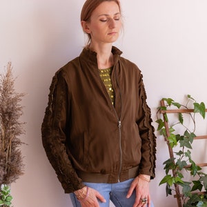 May include: A brown bomber jacket with ruffled sleeves. The jacket has a zipper closure and a  pocket on the left side. The model is wearing blue jeans.