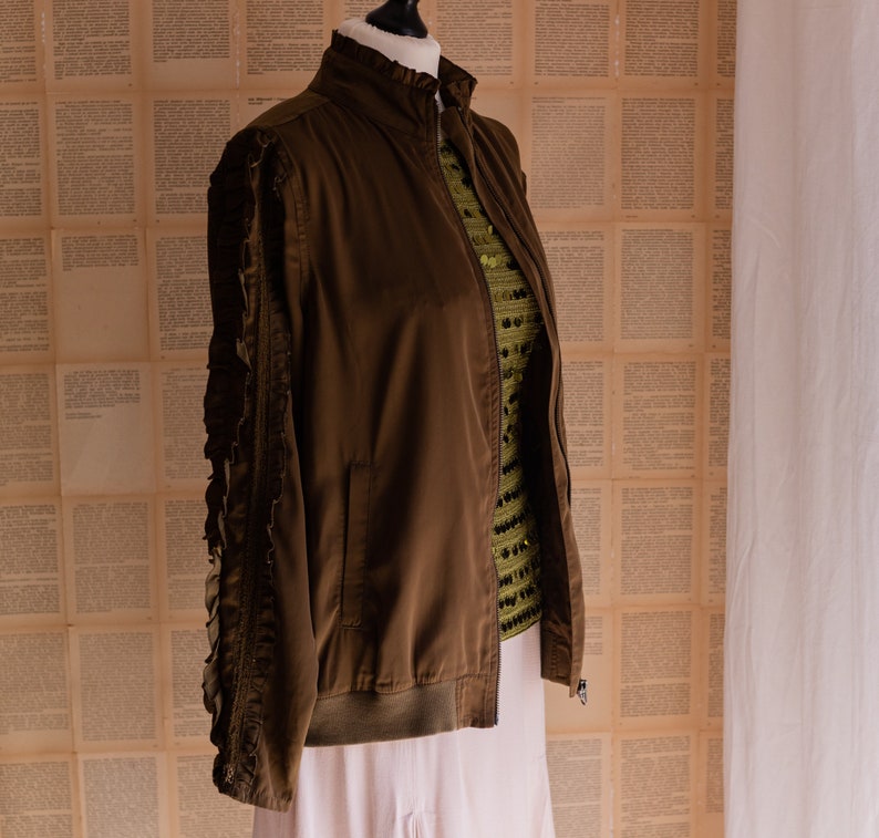 May include: A brown bomber jacket with ruffled sleeves. The jacket is on a mannequin and has a zipper closure.