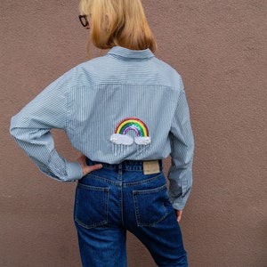 May include: A blue and white striped button-down shirt with a rainbow and cloud sequin patch on the back. The shirt is worn with blue jeans.