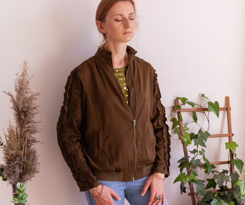 May include: A brown bomber jacket with ruffled sleeves. The jacket has a zipper closure and a  pocket on the left side. The model is wearing blue jeans.