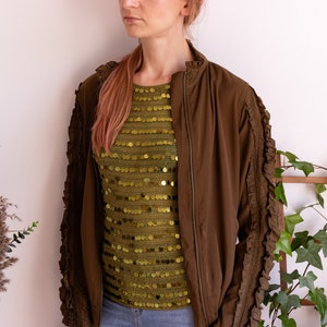May include: A brown bomber jacket with ruffled sleeves and a zipper closure. The jacket is worn over a green sequined top.