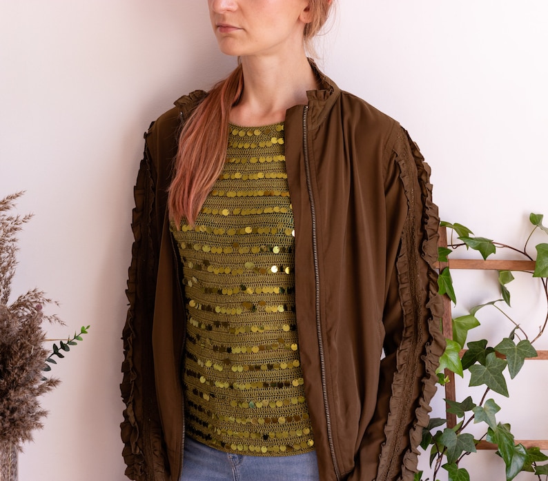 May include: A brown bomber jacket with ruffled sleeves and a zipper closure. The jacket is worn over a green sequined top.