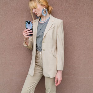 May include: A woman wearing a beige blazer with a light grey patterned shirt underneath. She is holding a black phone case with a cartoon character on it. She is wearing blue tassel earrings and a turquoise necklace.