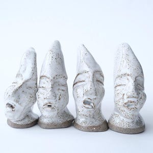 May include: Four white ceramic face sculptures with speckled details and a textured finish. Each sculpture has a unique expression, with closed eyes and open mouths. The bases are a neutral brown color, and the background is white.