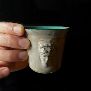 May include: A small ceramic mug with a teal blue rim and a raised design of a face on the side.