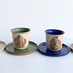 Może przedstawiać: Cztery ceramiczne filiżanki ze spodkami w różnych kolorach. Każda filiżanka posiada na froncie unikalny, wypukły relief przedstawiający twarz. Zestaw zawiera paletę kolorów kremowego, zielonego, niebieskiego i różowego z pasującymi spodkami.