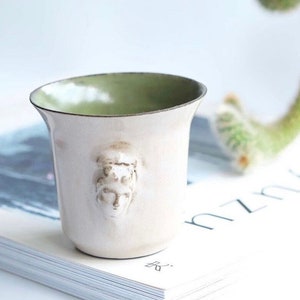 May include: A cream-colored ceramic cup with a green interior rim and a sculpted face design. The cup is placed on a book with the word "yuzu" visible. A cactus is in the background.