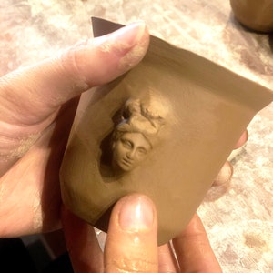 May include: A brown ceramic mug with a raised image of a woman's face on the front.
