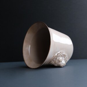 May include: A white ceramic mug with a lion's head sculpted on the side. The mug is tilted on its side, revealing the inside of the mug.