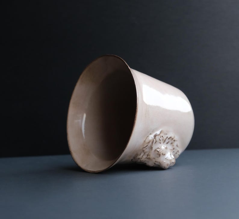 May include: A white ceramic mug with a lion's head sculpted on the side. The mug is tilted on its side, revealing the inside of the mug.