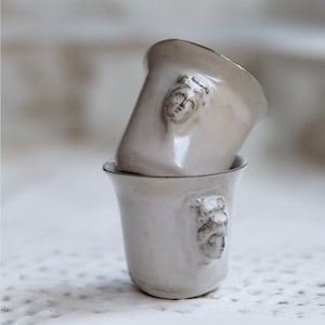 May include: Two white ceramic cups with a raised face design. The cups are stacked on top of each other.