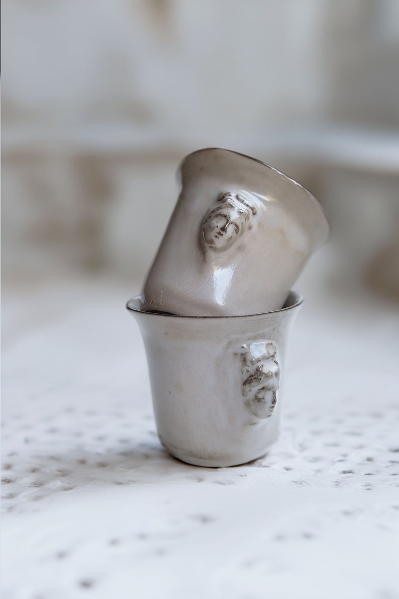 May include: Two white ceramic cups with a raised face design. The cups are stacked on top of each other.