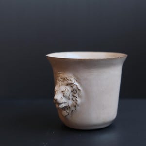 May include: A white ceramic cup with a lion's head sculpted on the side. The cup is sitting on a dark grey surface.