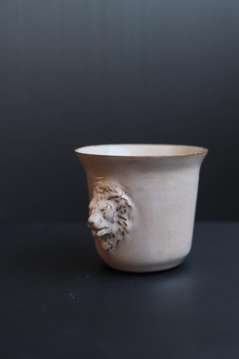 May include: A white ceramic cup with a lion's head sculpted on the side. The cup is sitting on a dark grey surface.