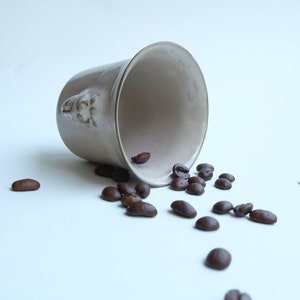 May include: A white ceramic mug with a face sculpted on the side, tipped over, spilling out dark brown coffee beans onto a white surface.