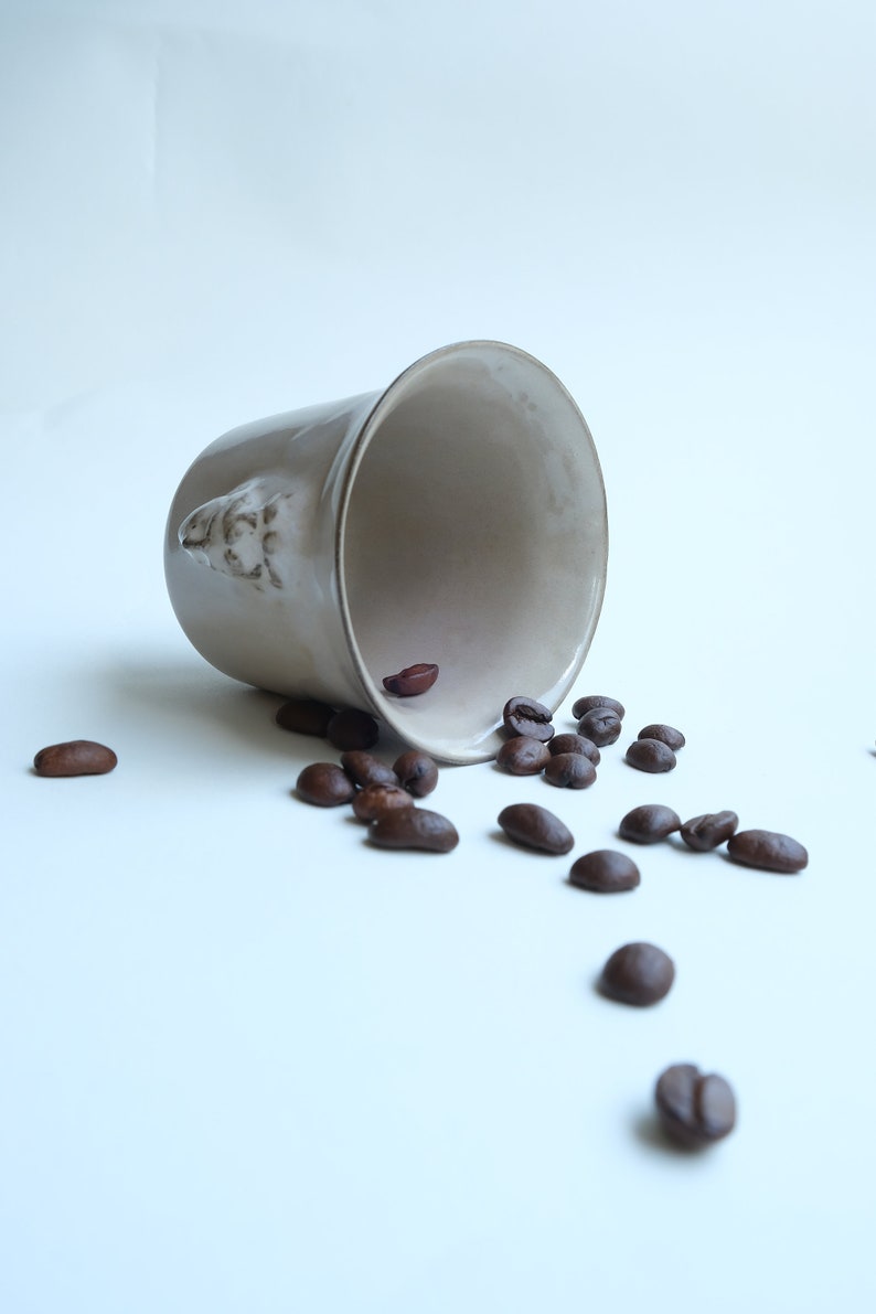 May include: A white ceramic mug with a face sculpted on the side, tipped over, spilling out dark brown coffee beans onto a white surface.