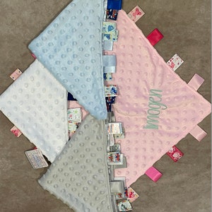 May include: Four baby security blankets in pastel colors: white, light blue, gray, and pink. Each blanket has a textured, dimpled surface and a row of colorful tags along one edge. The pink blanket has the name "Imogen" embroidered on it.