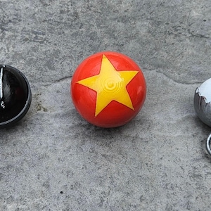 May include: Three small, round objects on a gray surface. A black shift knob with white markings, a red ball with a yellow star, and a silver-colored object with a white sphere inside.